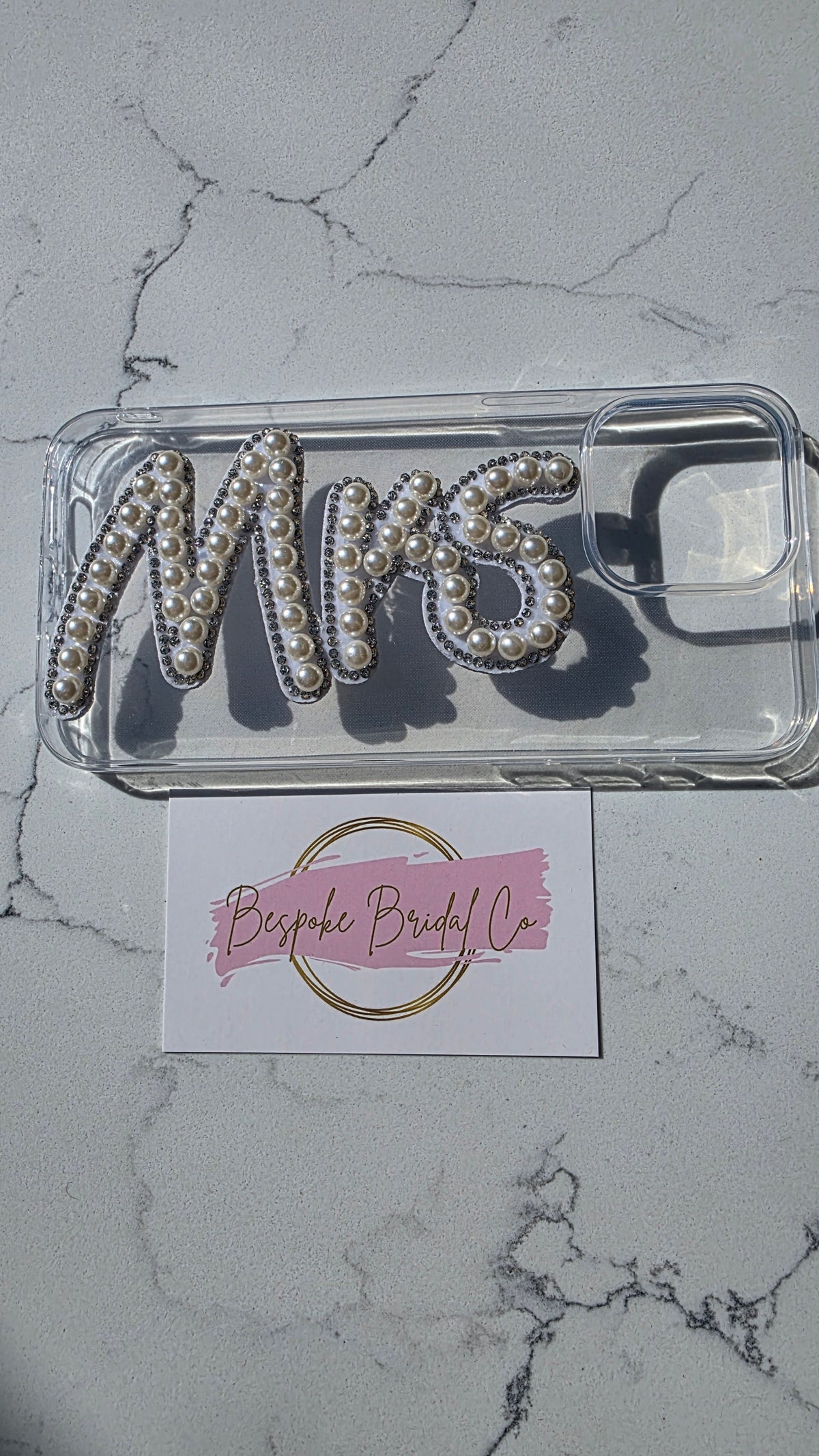 A clear phone case featuring the word 'MRS' embellished with white pearls and silver rhinestones, displayed on a light gray marble surface above a pink and white 'Bespoke Bridal Co' card.