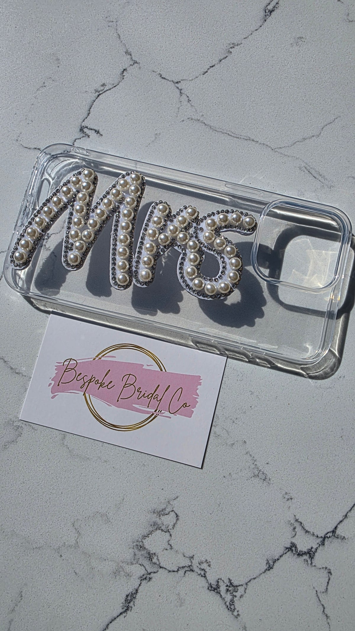 A clear phone case featuring the word 'MRS' in raised letters embellished with white faux pearls and silver rhinestones, displayed on a light gray marble surface next to a pink and white business card for 'Bespoke Bridal Co'.