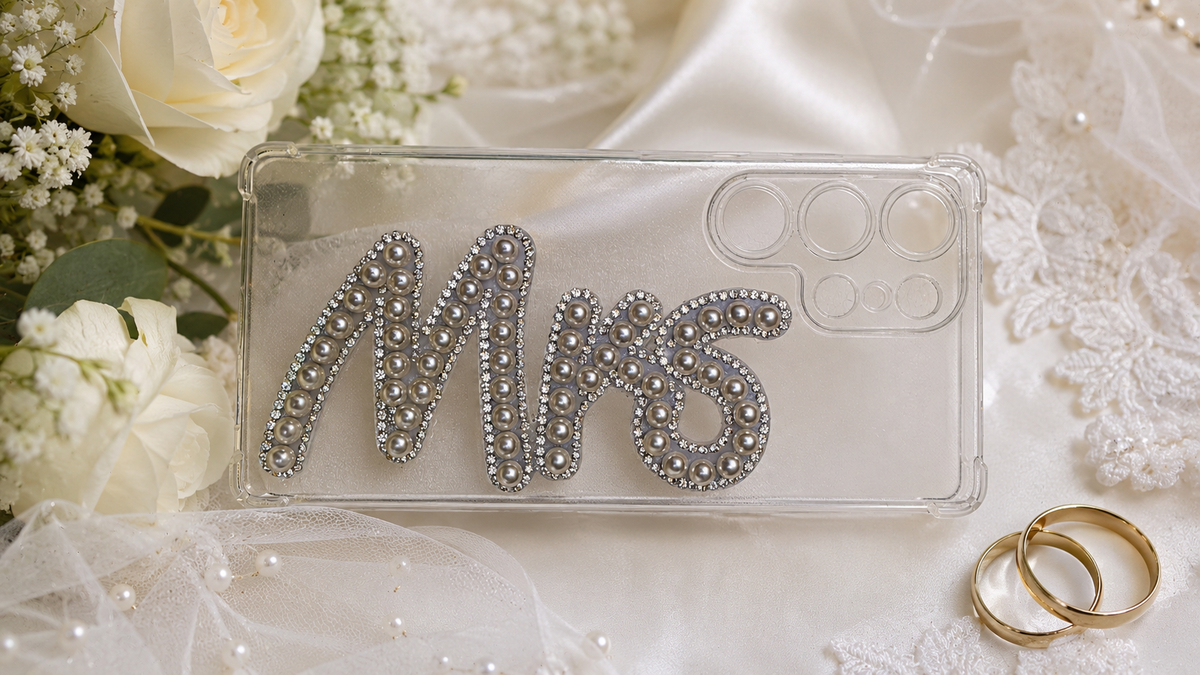 A clear phone case with the word 'MRS' spelled out in sparkling silver rhinestones, styled on a white satin fabric with lace, white roses, and two gold wedding rings.